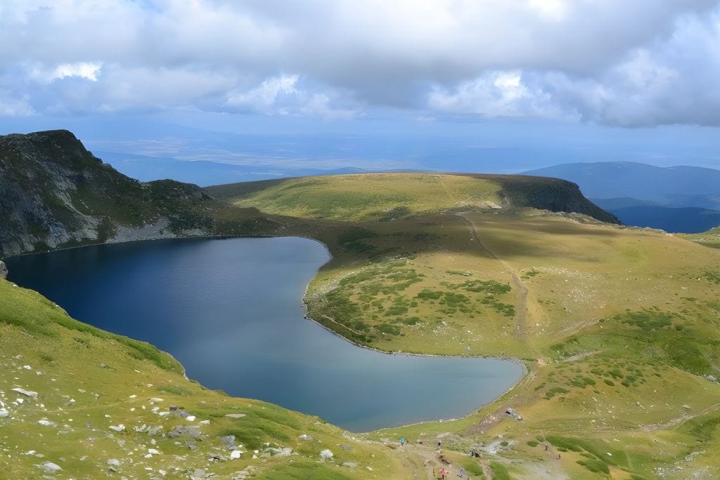 Rila Bulgaria Spiritual: The Most Spiritual Places to Visit in Bulgaria! A breathtaking view of Kidney Lake, one of the Seven Rila Lakes in Bulgaria, nestled among green hills and rocky terrain. The clear blue waters of the lake contrast with the surrounding grassy landscape, under a sky filled with dramatic clouds, creating a serene and picturesque alpine scene.