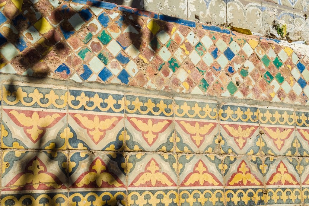 The Seven Saints of Marrakech (Local Insight of Marrakesh) A detailed view of a wall decorated with colorful geometric and floral-patterned tiles. The top section features small diamond-shaped tiles in shades of green, blue, yellow, and white, while the bottom section showcases larger tiles with intricate yellow and red floral motifs against a blue and beige background. Shadows cast by nearby objects add depth to the vibrant scene.