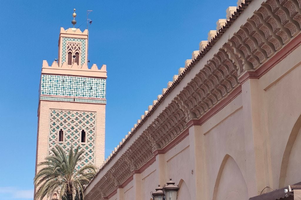 The Seven Saints of Marrakech (Local Insight of Marrakesh) A bustling market scene with vibrant stalls selling colorful clothing, jewelry, and souvenirs set against the backdrop of a mosque with intricate tilework and a tall minaret. The beige walls of the mosque are adorned with detailed carvings, and a palm tree adds a touch of greenery to the scene under a clear blue sky.