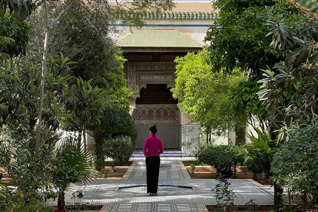 The Seven Saints of Marrakech (Local Insight of Marrakesh) A woman in a pink jacket and black pants stands at the end of a tiled pathway in a lush garden. The pathway leads to an ornate building entrance adorned with intricate designs and green tiles. Tall trees and dense foliage surround the serene garden scene under a partly cloudy sky.