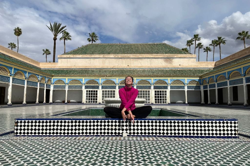 The Seven Saints of Marrakech (Local Insight of Marrakesh) A woman in a pink jacket and black pants sits smiling on the edge of a tiled fountain in the courtyard of a historic palace. The courtyard features a green-tiled roof and an arcade with blue and yellow accents, set against a backdrop of palm trees and a partly cloudy sky. The intricate tilework of the courtyard floor and fountain adds to the grandeur of the setting.
