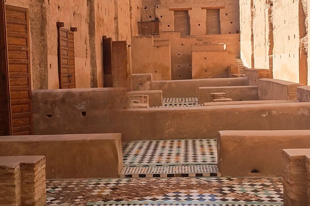 The Seven Saints of Marrakech (Local Insight of Marrakesh) A view of an ancient structure featuring weathered stone walls and intricate, colorful tile flooring. The walls have multiple wooden doors and small openings, while the floor displays a mix of geometric patterns. The earthy tones of the architecture contrast with the vibrant tiles, highlighting the historical and cultural significance of the site.