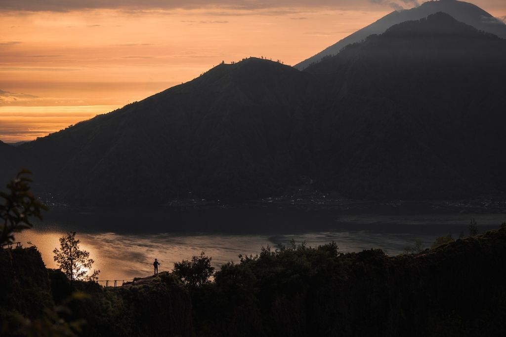 Mount Batur Sunrise Trekking: A Spiritual Journey A lone hiker stands on a ridge overlooking Mount Batur and its surrounding landscape at sunrise. The sky is painted with warm orange hues, casting a serene glow over the lake and the silhouetted mountains in the background. The scene evokes a sense of solitude and connection with nature.