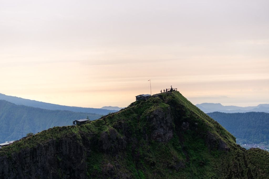 Mount Batur Sunrise Trekking: A Spiritual Journey Hikers stand at the summit of Mount Batur, silhouetted against the early morning sky. The scene captures the green, rugged peak with the vast landscape stretching into the distance, creating a sense of accomplishment and awe. The soft pastel colors of the dawn sky add to the tranquil atmosphere.