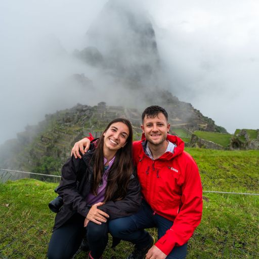Mount Batur Sunrise Trekking: A Spiritual Journey A smiling couple, Sara and Ryan, poses on a grassy hill at Machu Picchu, surrounded by mist and cloud-covered ruins in the background. The woman is wearing a black jacket and the man a red jacket, both appearing happy and adventurous.