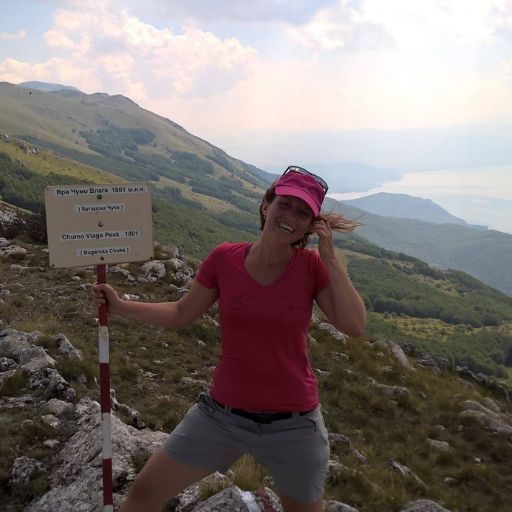 Rila Bulgaria Spiritual: The Most Spiritual Places to Visit in Bulgaria! Radina, a woman wearing a pink hat and shirt stands on a rocky hillside, smiling as she holds a sign that reads "Chumo Vlaga Peak 1801 m" in Macedonia. The background features a scenic view of rolling hills and a distant lake under a partly cloudy sky.