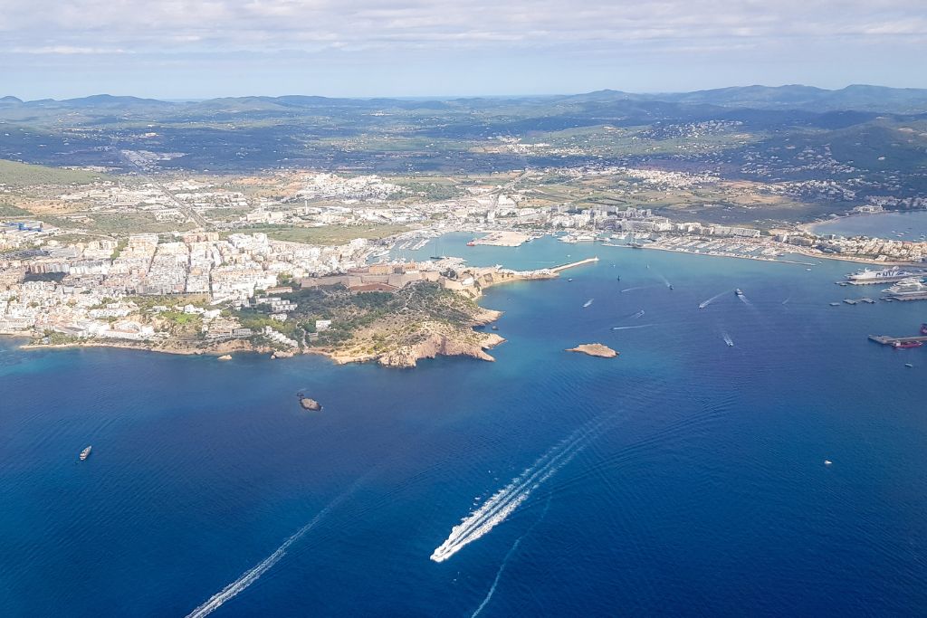 Aerial view of the coastal of Es Vedra’s Spiritual Significance with a sprawling urban area and a marina. Boats can be seen creating trails in the blue waters, and the cityscape is surrounded by green hills and mountains in the distance. The coastline features rocky promontories and clear, calm seas.