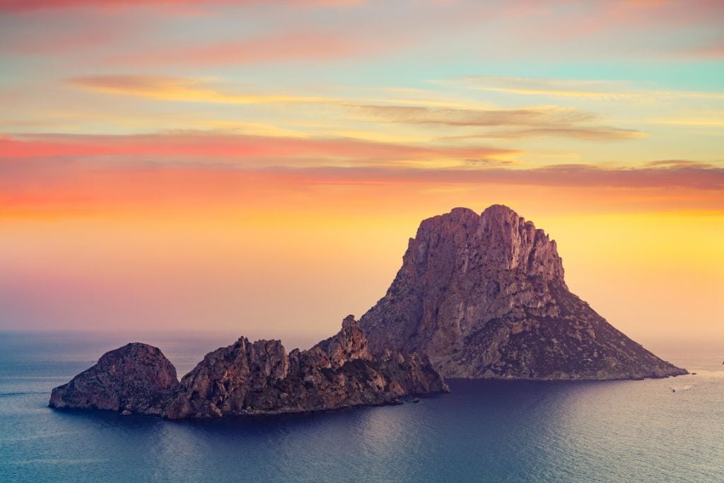 Rocky island formation in the sea at sunset, with the sky painted in vibrant hues of orange, pink, and yellow. The calm waters surround the rugged island, creating a serene and picturesque landscape.