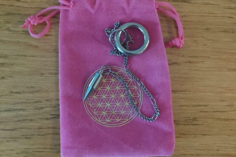 A silver pendulum with a conical-shaped weight and chain, placed on a pink drawstring pouch. The pouch has a geometric flower-like design printed in gold on the front, resting on a wooden surface.