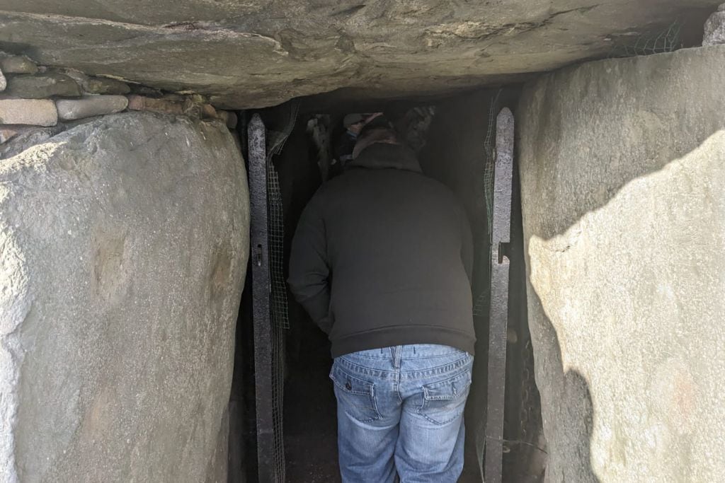 Visit Ireland's Greatest Megalithic Attraction: The Legendary Newgrange This image shows a person bending down to enter the narrow stone passageway of the Newgrange tomb. The entrance is framed by large stones, and the ceiling is low, requiring visitors to crouch as they make their way inside. The surrounding stones are weathered, adding to the ancient and mysterious atmosphere of the site.
