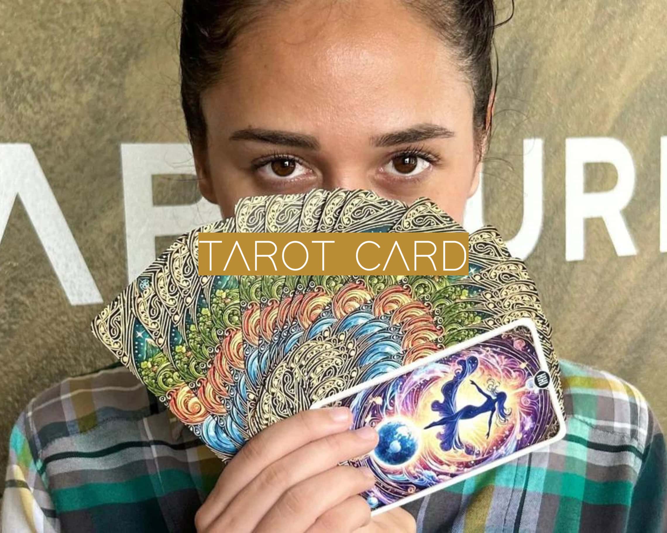 The image shows a person holding up a fan of intricately designed tarot cards, with one card featuring a cosmic, mystical illustration visible. The text "TAROT CARD" is overlaid in bold, stylized letters on a gold rectangle in the center of the image.