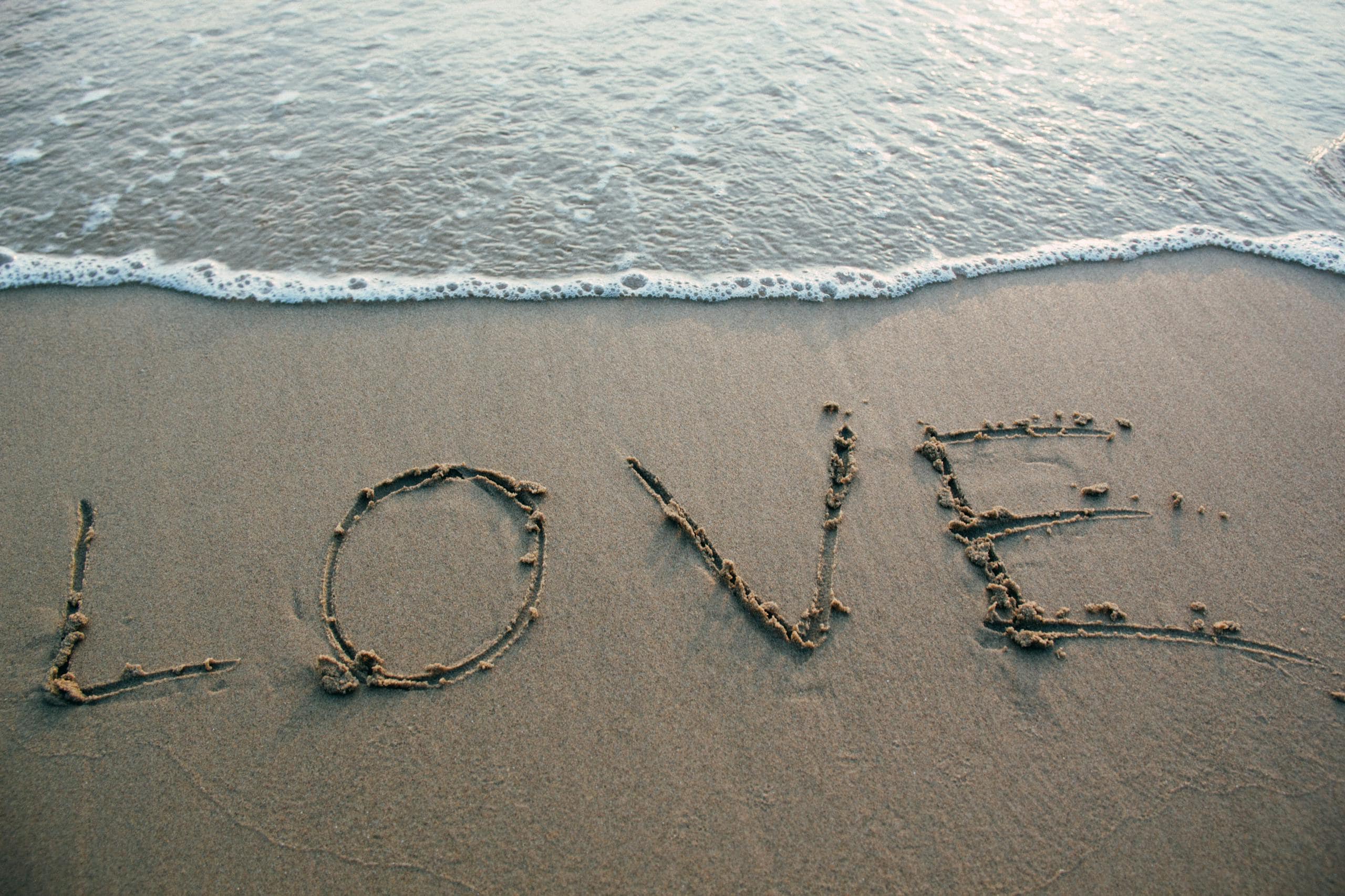 7744 Angel Number - Meaning in Love, Twin Flame, Career, and Health A heart symbol drawn in the wet sand on a seashore, with the word 'Love' written inside the heart. Gentle waves approach the shore, with soft brown sand beneath and a calming, romantic atmosphere showing 7744 Angel Number Love Meaning.