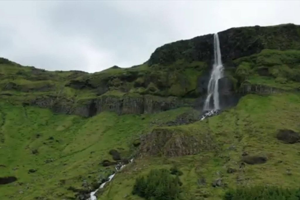 A tall, narrow waterfall cascades down a lush green hillside, surrounded by moss-covered rocks and rugged terrain. The water flows gently from the cliff's edge, creating a serene contrast against the vibrant greenery, under a cloudy sky that adds to the moody atmosphere of the landscape.