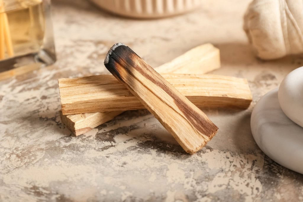 Palo Santo Vs Sage: Which One is Better? This image shows a few pieces of Palo Santo wood on a textured surface, with one of the pieces charred at the top. The background includes soft, neutral elements, such as smooth stones and a diffuser bottle, evoking a calming and natural ambiance commonly associated with aromatherapy or spiritual cleansing practices.