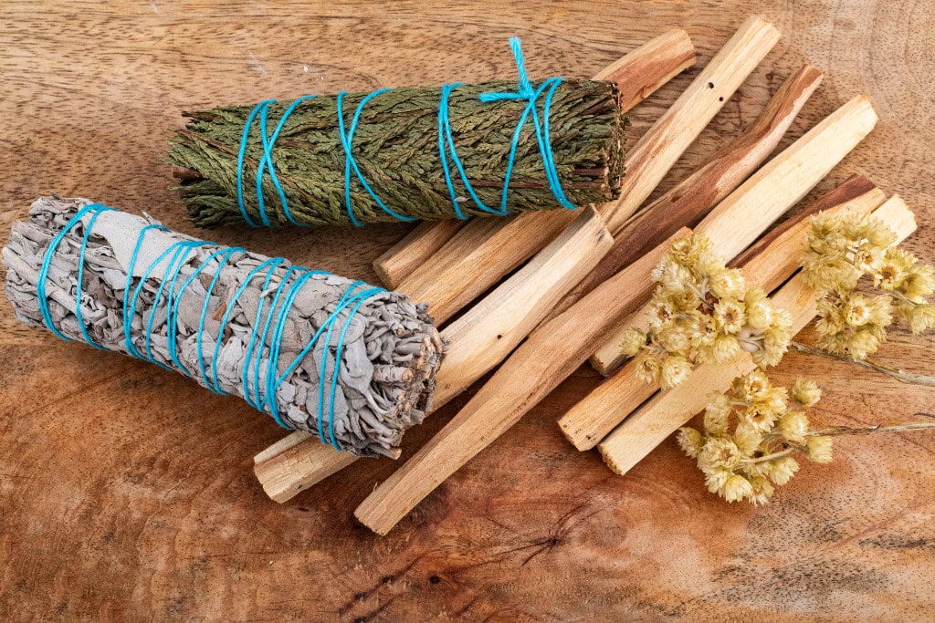 Palo Santo Vs Sage: Which One is Better? This image features a collection of spiritual cleansing tools, including two sage bundles wrapped in blue string, several pieces of Palo Santo wood, and a small cluster of dried flowers, all laid out on a wooden surface. The items are commonly used in smudging rituals and energy purification practices.