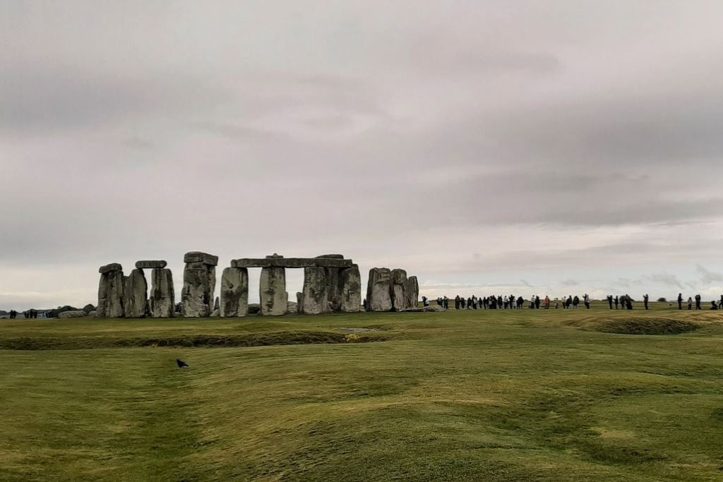 This image shows Stonehenge, the iconic prehistoric monument in England, with a cloudy sky overhead. The ancient stone circle is surrounded by a large grassy field, and a line of visitors can be seen in the distance walking around the monument. A single bird is visible in the foreground on the grass.