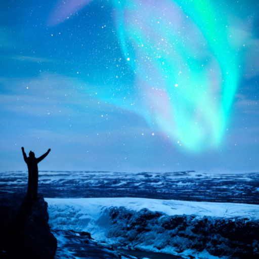A silhouetted person stands with arms raised in awe beneath a vibrant display of the Northern Lights in a snow-covered landscape. Shades of turquoise and pink illuminate the night sky, casting a magical glow over the expansive, icy terrain below. The image captures a moment of connection with nature's beauty and wonder.