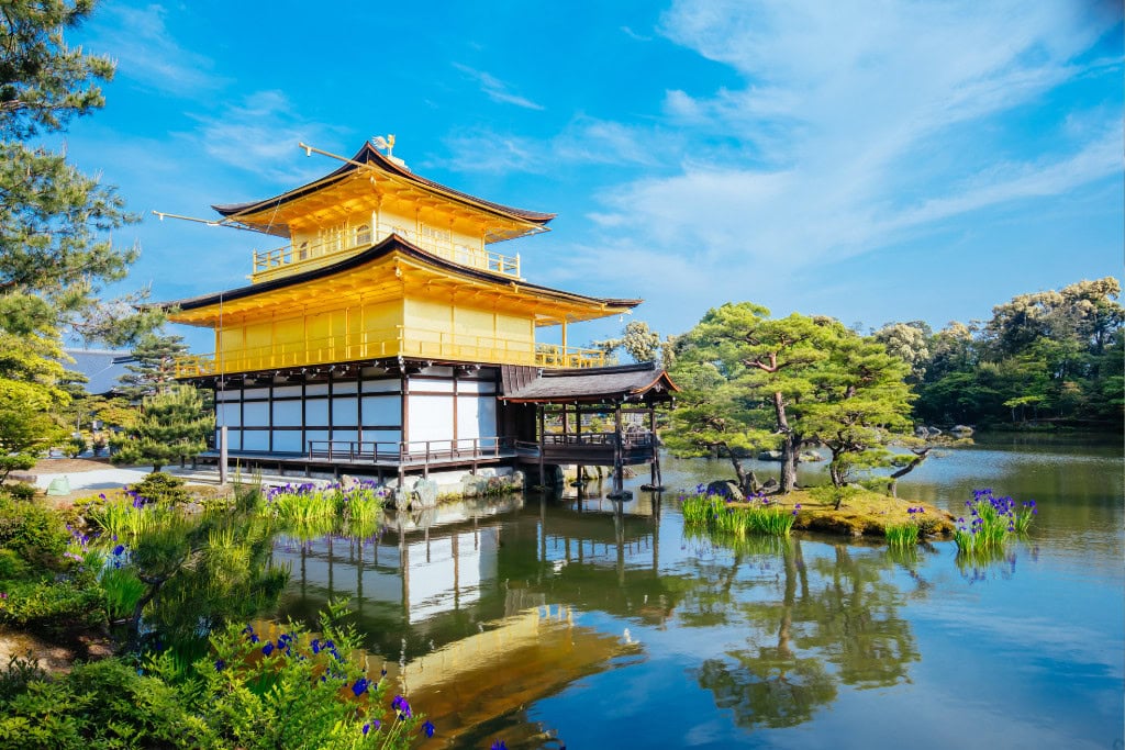 Historical Sites in Kyoto (Guide Insight Kyoto's Landmarks) The image showcases the famous Golden Pavilion, or Kinkaku-ji, in Kyoto, Japan, reflecting beautifully on the surrounding pond under a clear blue sky. The golden exterior shines brightly amid lush green trees and vibrant plants, creating a serene and picturesque view of one of Kyoto’s most iconic historical sites.