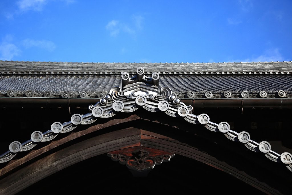 Historical Sites in Kyoto (Guide Insight Kyoto's Landmarks) The image shows a close-up of a traditional Japanese temple roof, with intricate tiles and ornate details set against a clear blue sky. The curved eaves are decorated with circular tiles, each featuring Japanese kanji symbols, reflecting the architectural elegance of historical sites in Kyoto. This craftsmanship highlights the cultural heritage and meticulous design common in Japanese temples.