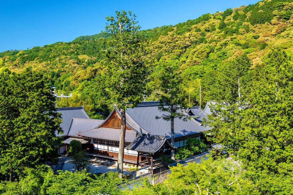 Historical Sites in Kyoto (Guide Insight Kyoto's Landmarks) The image depicts a traditional Japanese temple complex nestled within a lush green forest, with rolling hills in the background under a bright blue sky. The rooftops of the wooden structures blend harmoniously with the surrounding natural landscape, reflecting the serene integration of architecture and nature typical of Kyoto’s historical sites. This peaceful setting exemplifies the tranquil beauty of Kyoto's countryside temples.