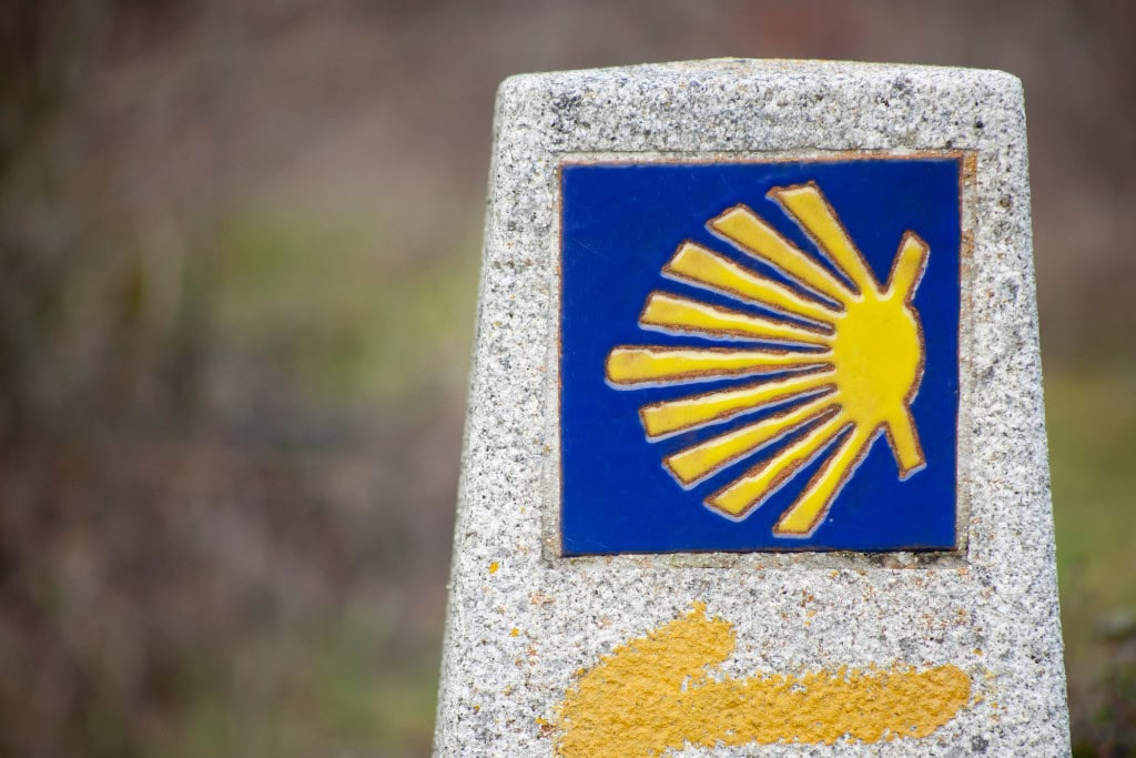 This image shows a close-up of a stone marker with the blue and yellow scallop shell symbol, a recognizable sign of the Camino de Santiago pilgrimage. Below it, a painted yellow arrow indicates the direction for travelers. The blurred background suggests a natural outdoor setting, emphasizing the path's journey and guidance for pilgrims.






