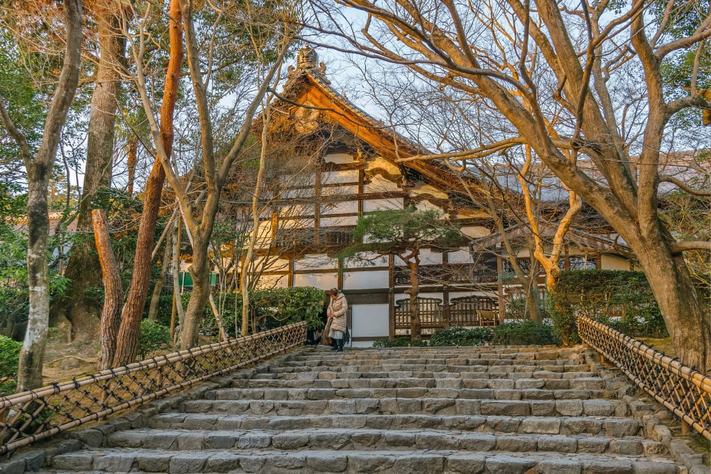 Historical Sites in Kyoto (Guide Insight Kyoto's Landmarks) The image shows the stone steps leading up to a traditional Japanese temple building, framed by bare trees, suggesting late autumn or early winter. The wooden structure with a steep, ornate roof is set amid a tranquil natural setting, with soft sunlight casting a warm glow. This pathway and architecture capture the quiet beauty and historic atmosphere of Kyoto's temples.
