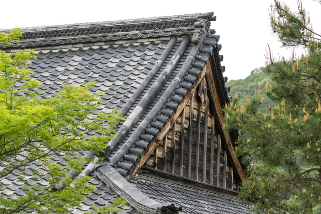 Historical Sites in Kyoto (Guide Insight Kyoto's Landmarks) The image highlights the detailed roof of a traditional Japanese temple with gray, intricately tiled shingles and ornamental ridges. The wooden structure beneath the roof shows signs of age, adding character, while the surrounding green foliage contrasts with the dark roof, creating a harmonious blend of architecture and nature typical of Kyoto’s historic sites.