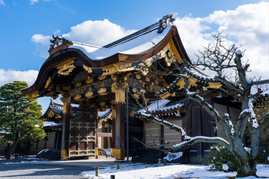 Historical Sites in Kyoto (Guide Insight Kyoto's Landmarks) The image shows the elaborate entrance gate of Nijo Castle in Kyoto, Japan, adorned with intricate gold detailing and traditional curved eaves, partially covered with a dusting of snow. The bright blue sky and surrounding trees create a serene winter scene, highlighting the castle's historical grandeur and seasonal beauty. This gate exemplifies Kyoto's rich architectural heritage and cultural significance.