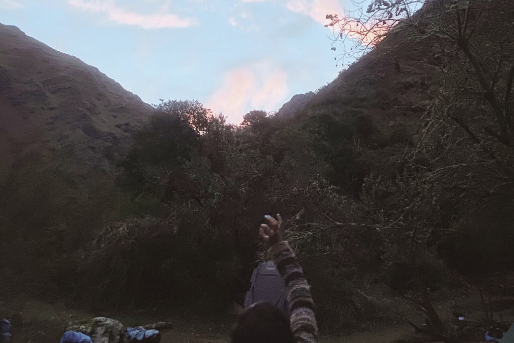 Spiritual Awakening: What’s It and How It Feels Like A person raises their arm in a gesture that evokes a sense of connection and spiritual awakening, set against a backdrop of rugged hills and a sky tinged with soft pink and blue hues at dawn or dusk. The scene is surrounded by natural foliage, adding to the tranquil and reflective atmosphere.