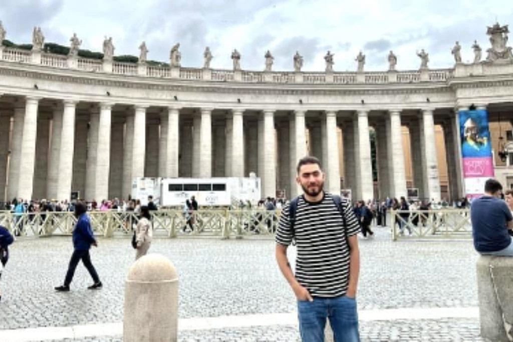 A man in a striped shirt standing in front of the iconic colonnades of St. Peter's Square in Vatican City, with a backdrop of statues, bustling visitors, and cloudy skies.