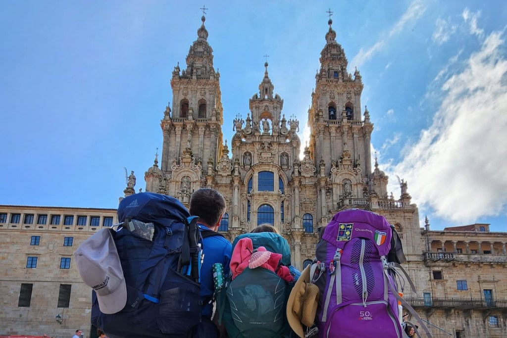 Three pilgrims with large backpacks stand in front of the Cathedral of Santiago de Compostela, gazing up at its ornate towers and intricate facade, a symbol of the spiritual journey of the Camino de Santiago.