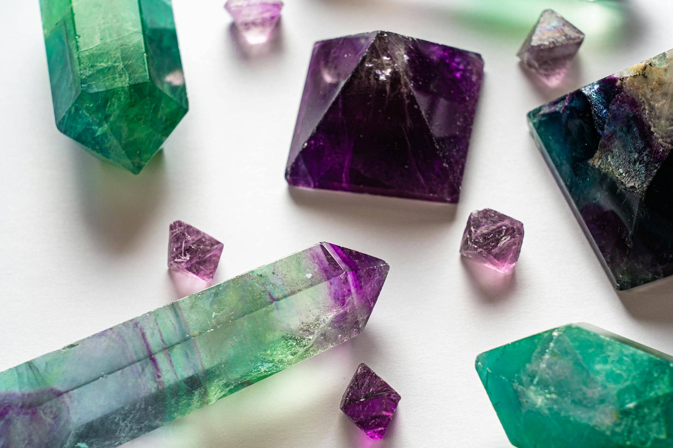 Close-up image of vibrant fluorite and amethyst gemstones displayed on a neutral surface. The fluorite stones feature a range of colors, including green, purple, and clear hues, with their smooth and polished textures catching the light. The amethyst stones showcase deep purple tones with crystalline facets that sparkle. 