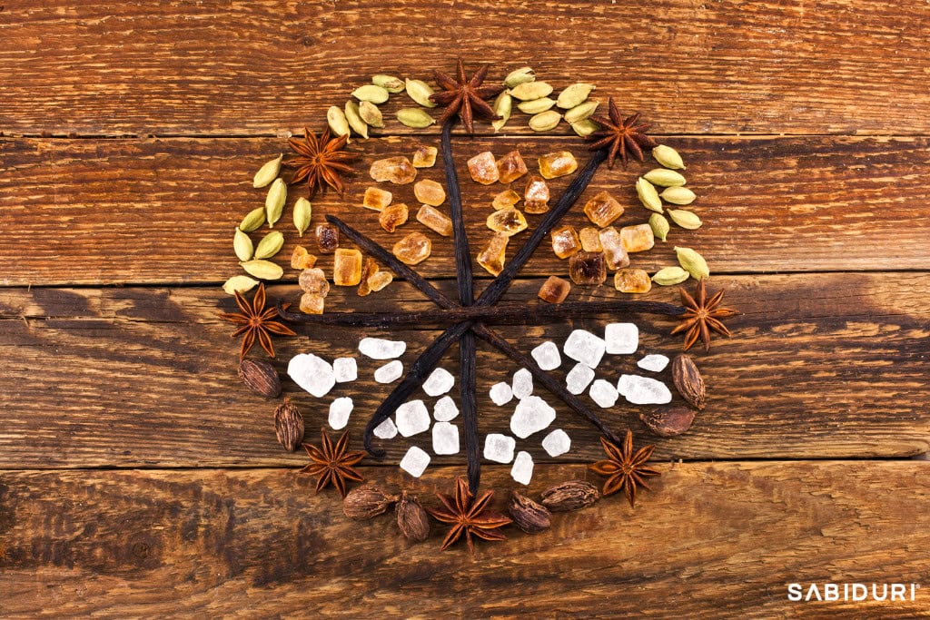A beautifully arranged Celtic Wheel made from natural spices and ingredients on a rustic wooden background. The design features sections divided by vanilla beans, with each quadrant filled with different elements: green cardamom pods, golden rock sugar, white sugar crystals, star anise, and black cardamom. The earthy tones and organic materials create a warm, nature-inspired aesthetic.