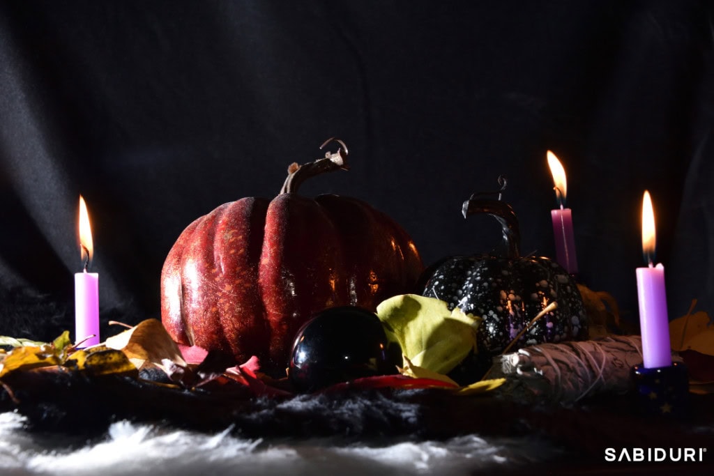 A Samhain Celtic Wheel altar setup featuring deep red and black pumpkins, four glowing purple candles, dried autumn leaves, a black crystal sphere, and a sage bundle on a dark, mystical backdrop. The scene embodies the spirit of Samhain, a sacred Celtic festival marking the end of the harvest and the thinning of the veil between worlds. The flickering candlelight enhances the ritualistic and mystical ambiance.