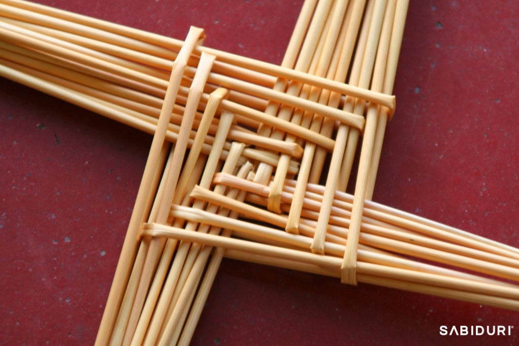 A close-up of a traditional Brigid’s Cross, woven from straw, symbolizing Imbolc on the Celtic Wheel. The intricate handwoven design represents protection and renewal, marking the transition from winter to spring in the ancient Celtic festival. The cross is set against a red background, emphasizing its earthy, natural texture and deep cultural significance.