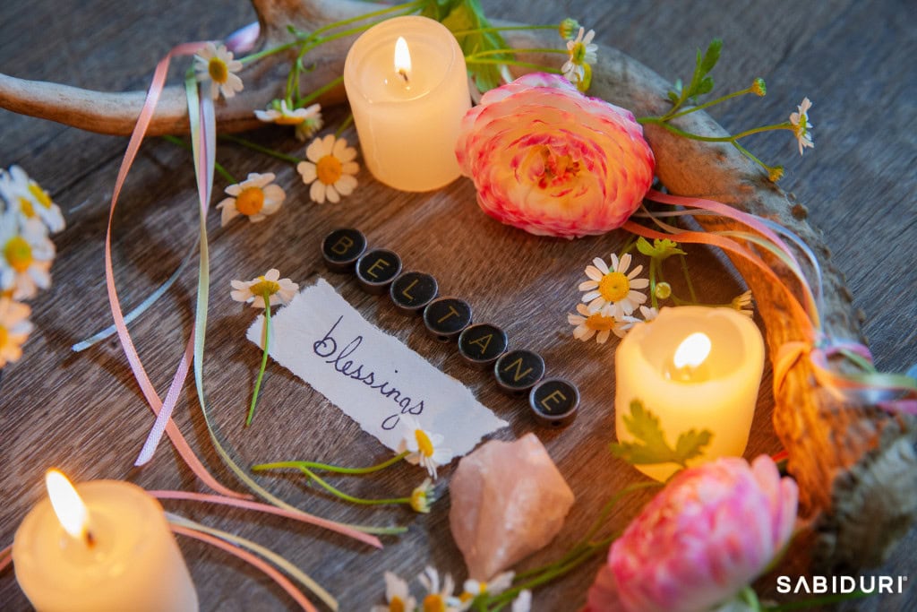 A beautifully arranged Beltane altar celebrating the Celtic Wheel, featuring glowing candles, fresh chamomile flowers, pink ranunculus blooms, and a handwritten note reading 'Beltane Blessings.' Delicate ribbons drape over a rustic antler, symbolizing fertility, passion, and the vibrant energy of spring. The warm candlelight enhances the sacred, joyful essence of this ancient Celtic fire festival.