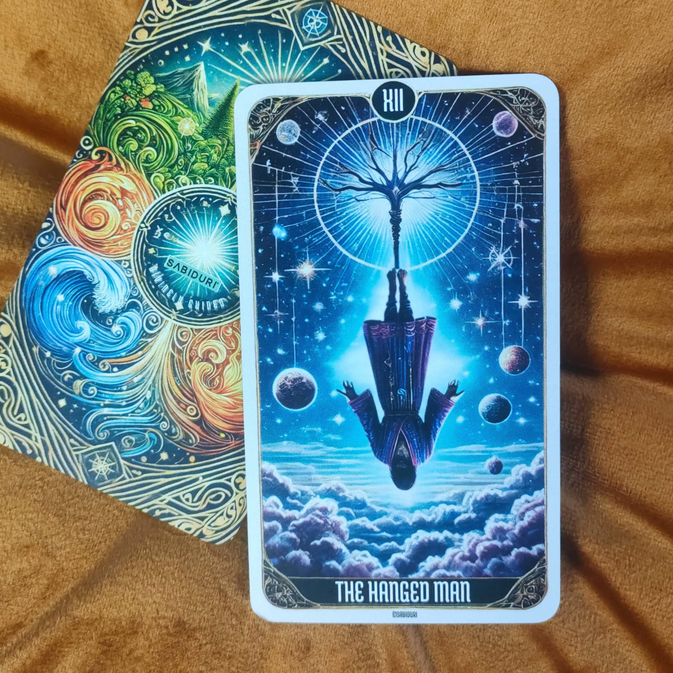 Tarot Card Meanings List: All Complete 78 Cards The card features a figure hanging upside down from a tree, with a cosmic scene in the background. The image suggests sacrifice, a change in perspective, and contemplation.