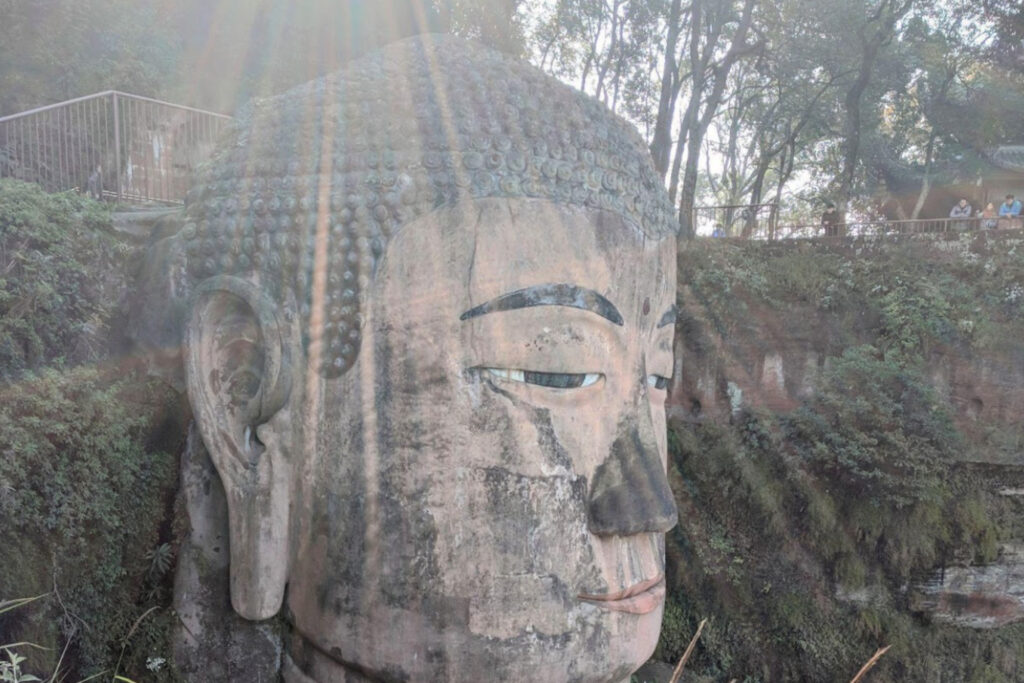 The Leshan Sprit Guide to the Leshan Giant Buddha Close-up of the massive stone head of the Leshan Giant Buddha in Sichuan, China, with sunlight streaming across the sculpture. The serene expression, detailed curls on the head, and weathered stone surface are framed by greenery and a viewing platform with onlookers in the background.