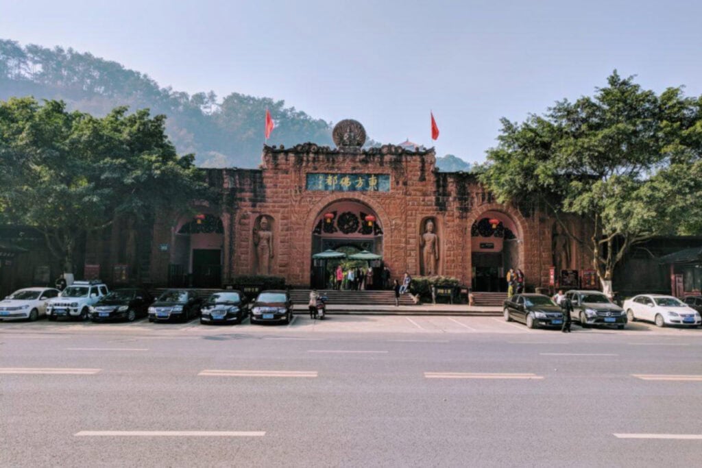 The Leshan Sprit Guide to the Leshan Giant Buddha Front view of the entrance to the Leshan Giant Buddha scenic area in Sichuan, China, featuring a grand reddish-brown stone facade with arched doorways, carved statues, and Chinese signage above the central arch. Several cars are parked in front, and visitors are walking up the steps, flanked by trees and mountains in the background.