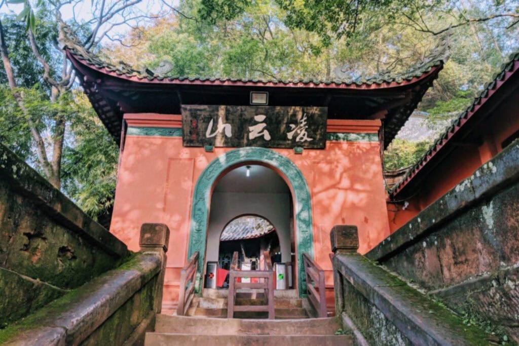 The Leshan Sprit Guide to the Leshan Giant Buddha Entrance to a traditional Chinese temple or pavilion with a curved tiled roof and ornate dragon carvings on the roof corners. The structure is painted coral-pink with teal trim around the arched doorway, and a wooden sign with Chinese characters hangs above. Stone steps and railings lead up through a lush, tree-covered area.