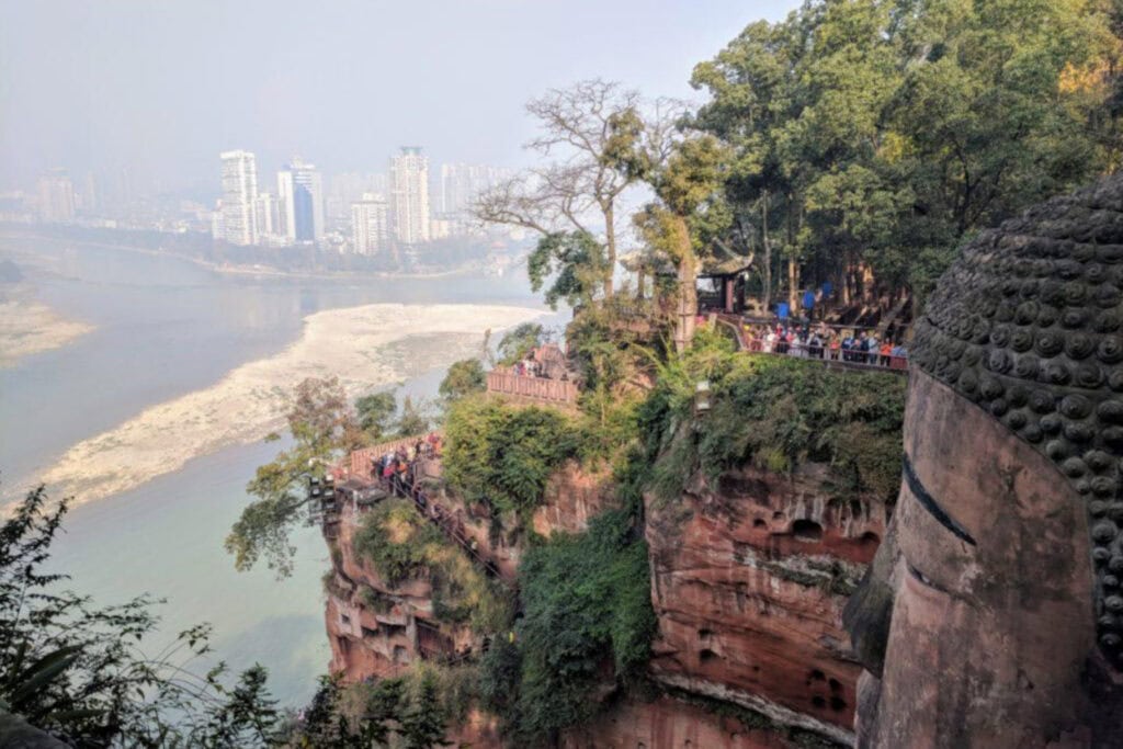 The Leshan Sprit Guide to the Leshan Giant Buddha View from above the Leshan Giant Buddha, showing the statue’s massive head in the foreground and crowds of tourists along cliffside walkways surrounded by trees. In the distance, the Dadu River winds past sandy banks, with the modern skyline of Leshan city rising through the haze.