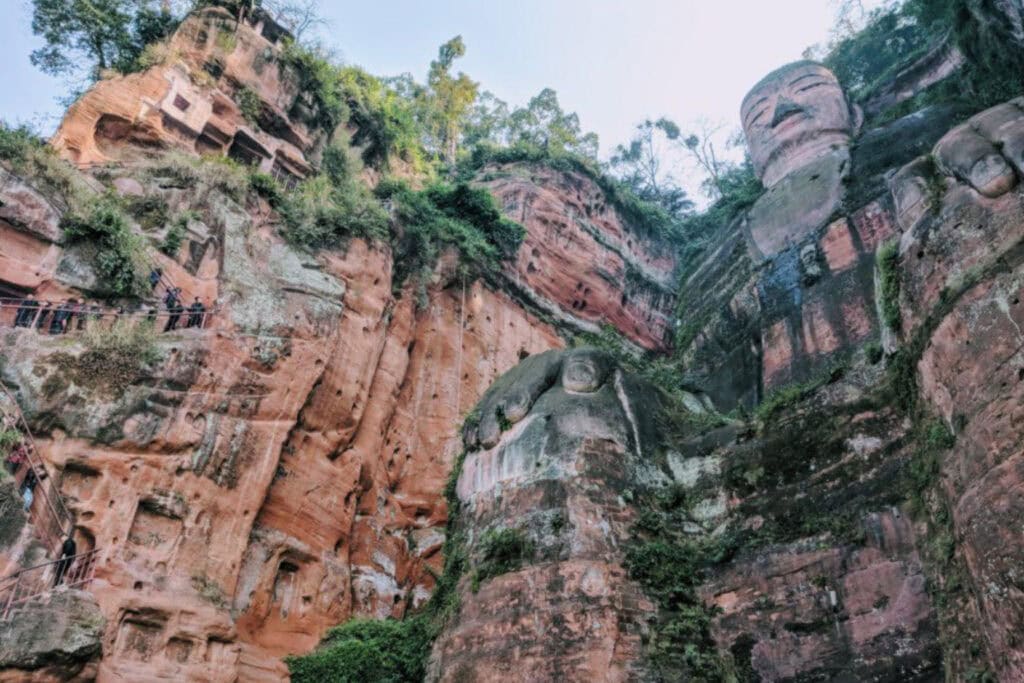 The Leshan Sprit Guide to the Leshan Giant Buddha Upward view of the towering Leshan Giant Buddha carved into a red sandstone cliff, showing its full body including massive hands resting on its knees. Tourists walk along narrow cliffside pathways, dwarfed by the sheer scale of the statue and the rugged, vegetation-dotted rock face.