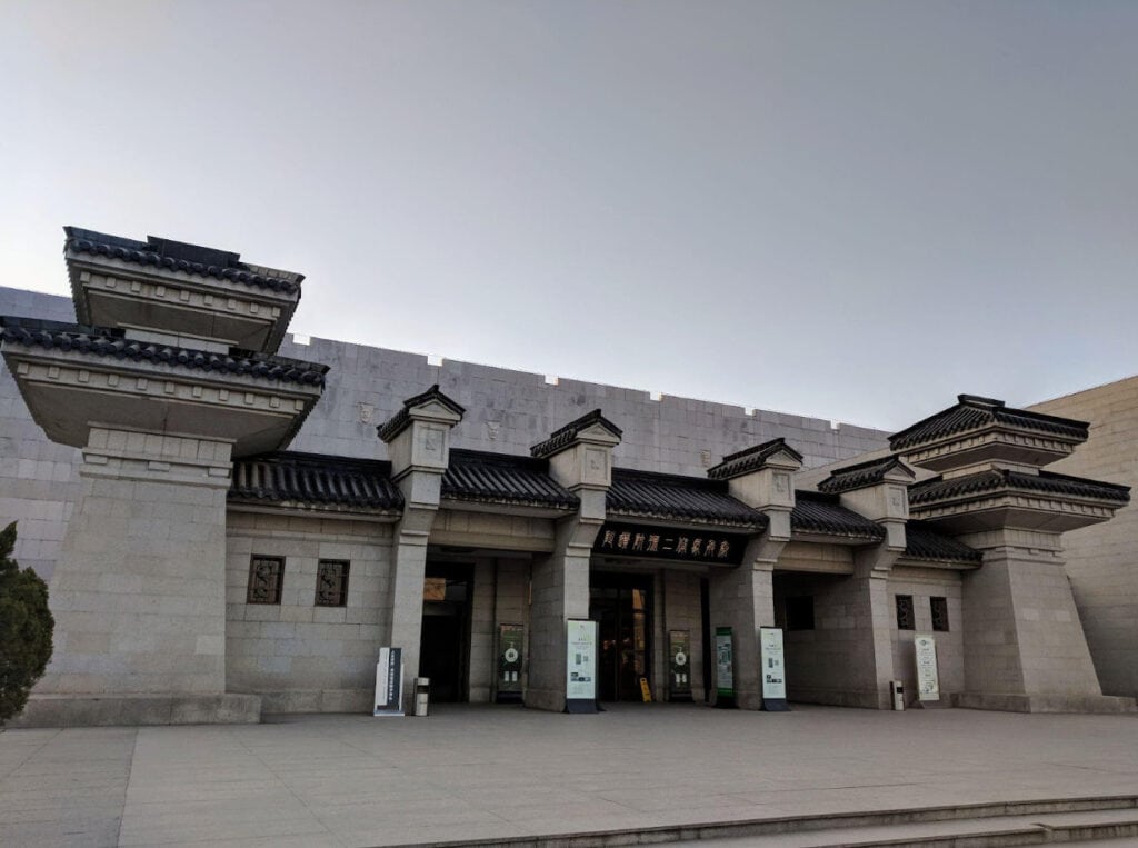 Xi'an Terracotta Warriors: History, Significance, and Travel Tips Exterior of the museum entrance at the Terracotta Warriors site in Xi’an, China, featuring a traditional Chinese architectural design with gray stone walls and tiered, dark-tiled roofs. The main entrance has signage in Chinese characters and is flanked by informational boards for visitors.