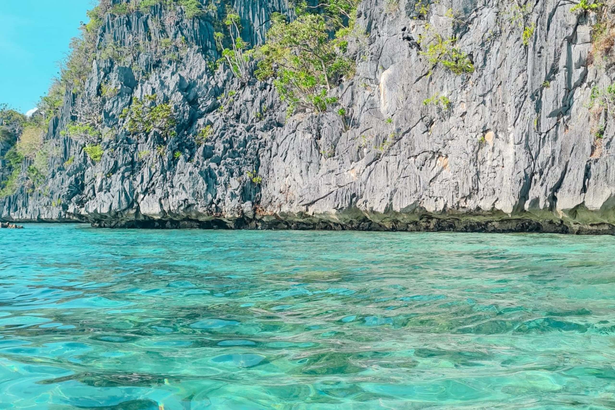 Crystal-clear turquoise waters with gentle ripples in Coron Palawan, Philippines. In the background, tall limestone cliffs rise dramatically, partially covered with green tropical vegetation. The sunlight illuminates the water, showcasing its stunning clarity and vibrant colors. This scene represents the natural beauty and iconic landscapes travelers experience during a 3-day trip to Coron Palawan, combining pristine nature, clear waters, and towering rock formations.
