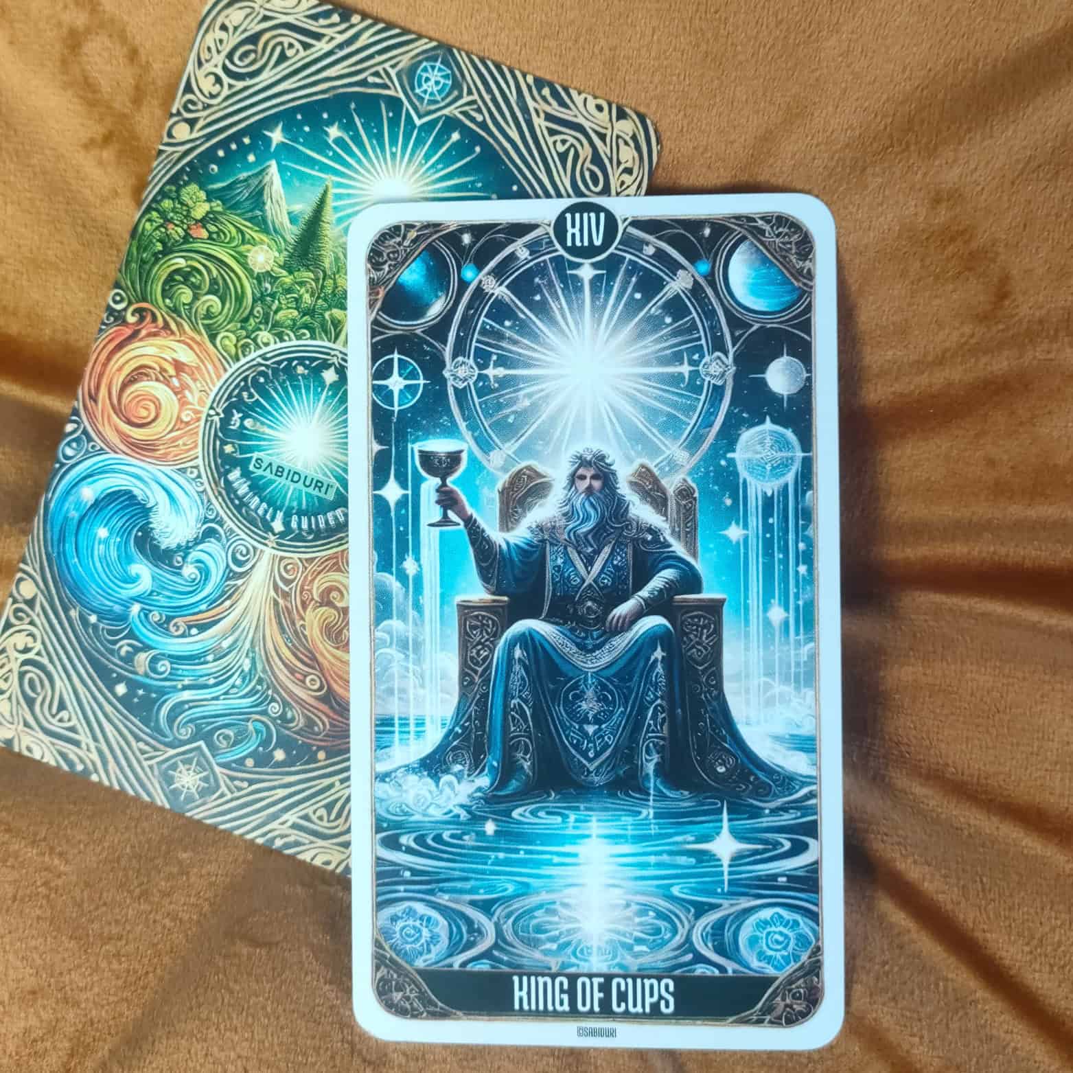 The King of Cups tarot card shown upright on a soft brown fabric background. The artwork features a regal king holding a cup, seated on a throne above turbulent waters with cosmic symbols behind him. In its upright position, the card represents emotional mastery, compassion, and wisdom.