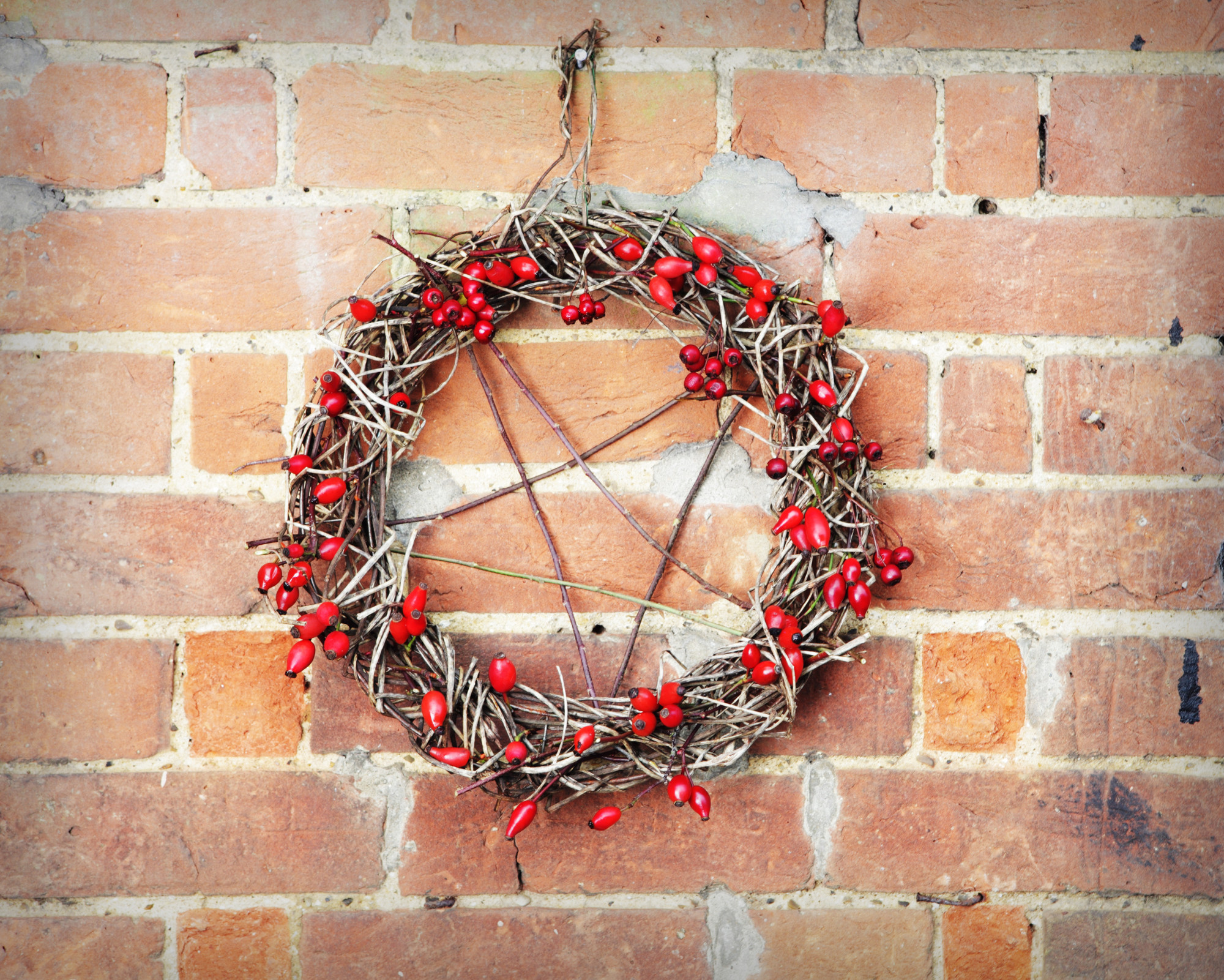 How to Celebrate Yule? Bring Yule Winter Solstice Into Your Life! A natural Yule wreath made of twigs and red berries hanging on a brick wall, symbolizing protection and winter magic for those exploring how to celebrate Yule.