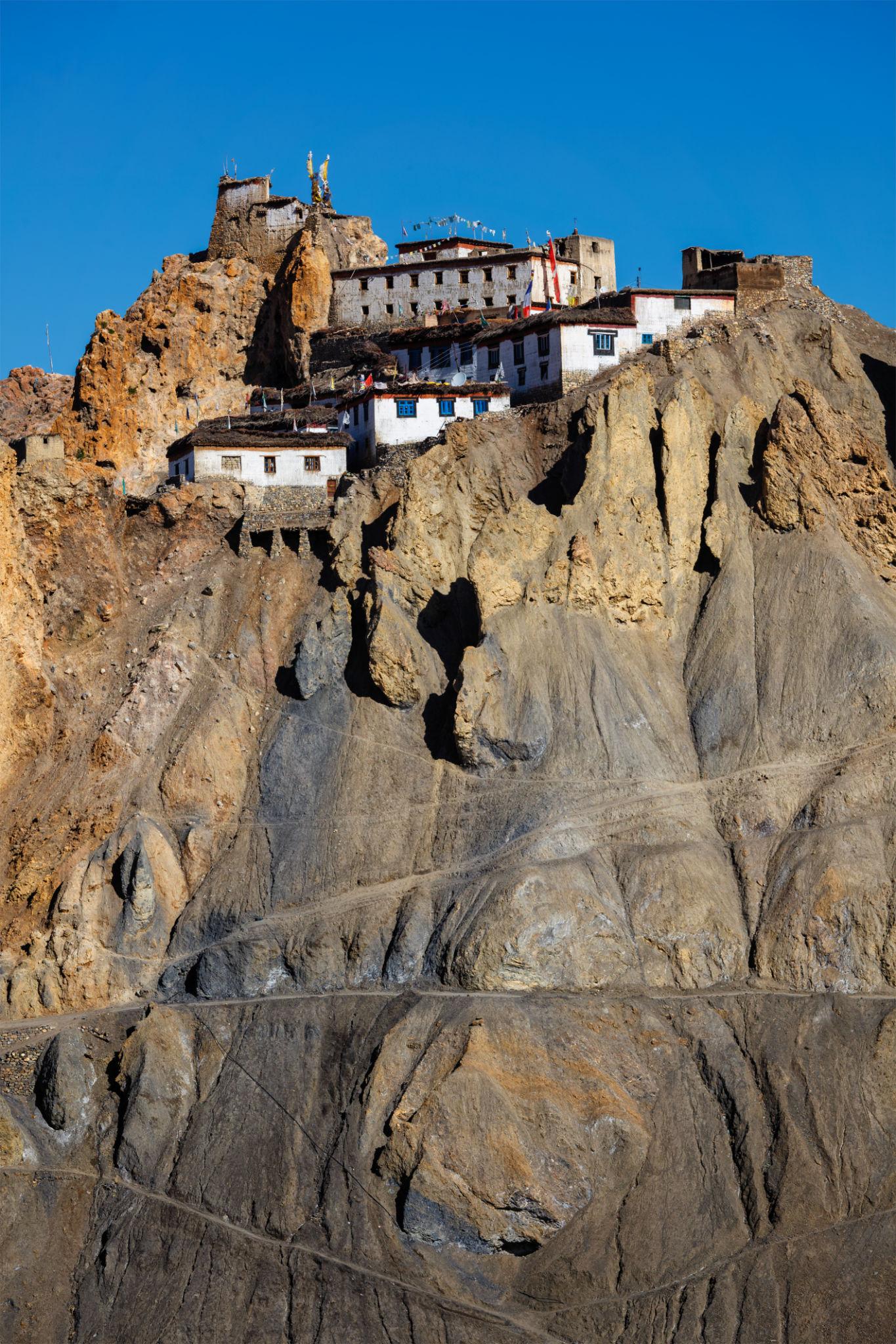 Dhankar Monastery perched dramatically on a rugged cliff in Spiti Valley, its whitewashed buildings rising above steep rock formations under a deep blue sky.