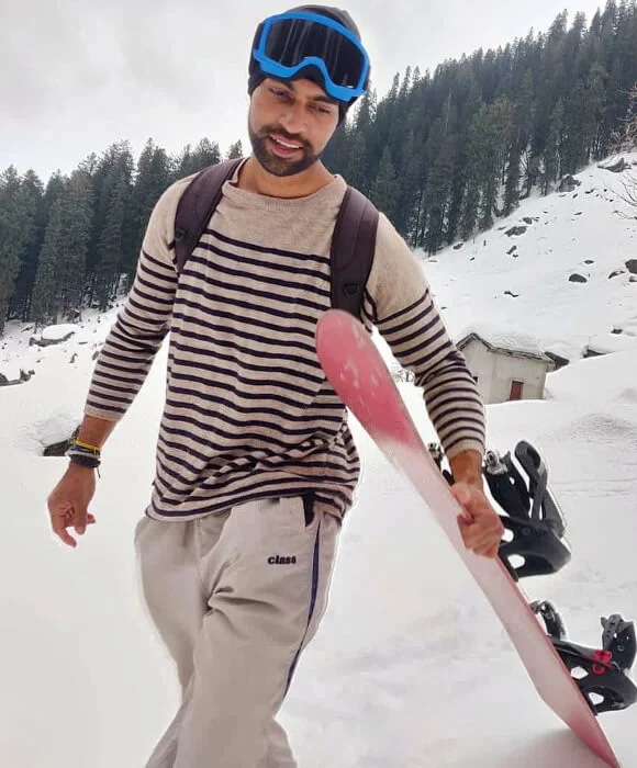 The Silence Between the Monasteries: What Spiti Valley Taught Me About Stillness Man wearing snow goggles and a striped sweater walking through a snowy mountain landscape while carrying a snowboard, with pine-covered hills and a small cabin in the background.