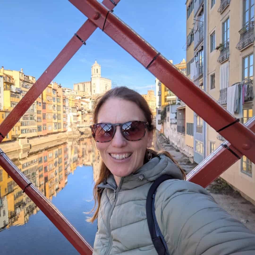Spanish Food Culture: Discover Spain Through Its Traditional Cuisine A person wearing sunglasses and a light green jacket smiles for a selfie while standing on a red iron bridge above a calm river. In the background, colorful historic buildings line the waterfront, reflecting in the water below, while a church tower rises above the city skyline. The scene captures the charm and beauty of Girona, Spain, on a clear sunny day.