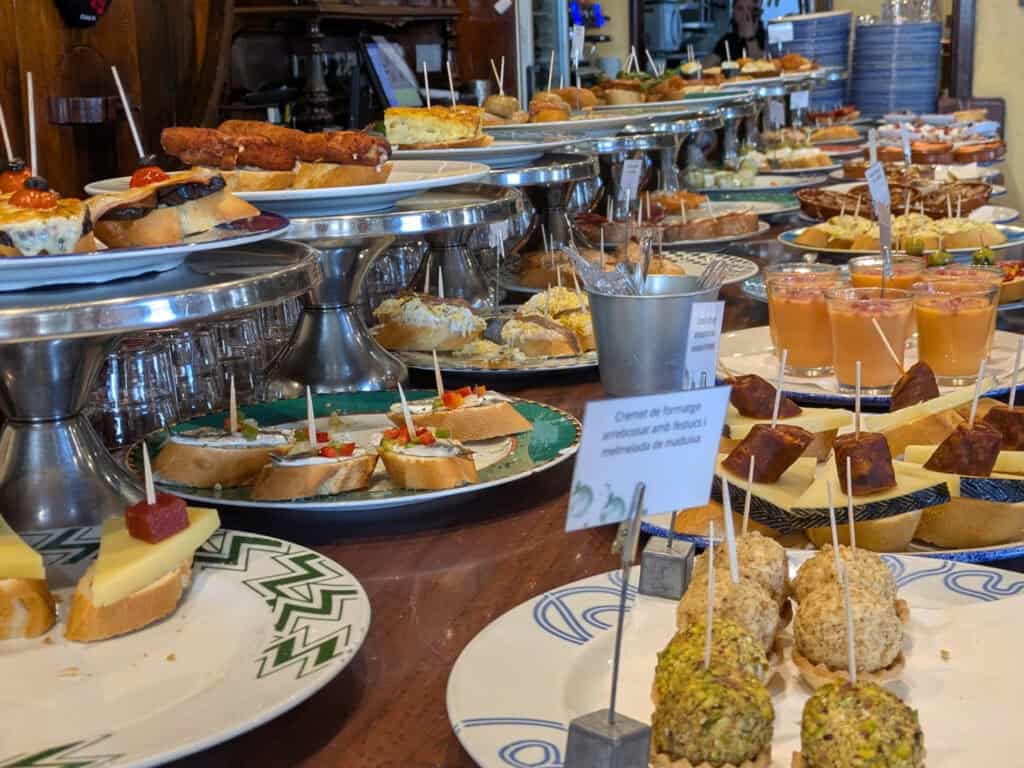 Spanish Food Culture: Discover Spain Through Its Traditional Cuisine A bustling Spanish tapas bar displaying dozens of pintxos and small plates on elevated trays and serving platters. The counter is filled with a variety of appetizers, including croquettes, cheese-topped bread, seafood bites, cured meats, and colorful finger foods secured with toothpicks. The warm atmosphere and abundance of dishes highlight the social dining traditions of Spanish food culture.